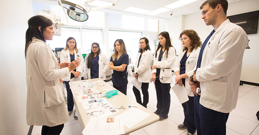 "UNT Health Physician Assistant students in white lab coats gathered around a table in a clinical setting, attentively listening to an instructor during a hands-on training session.