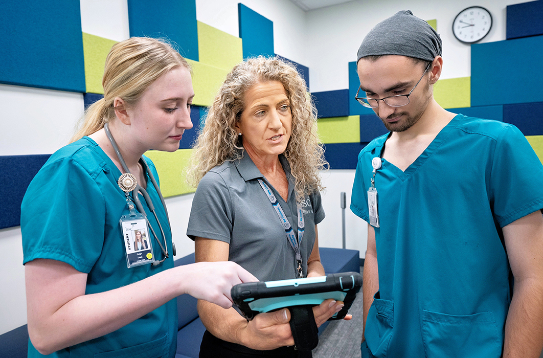 Two students in teal scrubs, on professor in Gray, looking over a tablet.